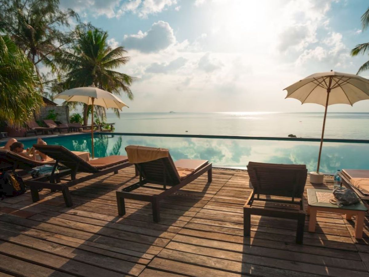 A tropical poolside scene with lounge chairs, umbrellas, and lush palm trees, overlooking a serene body of water under a partly cloudy sky.