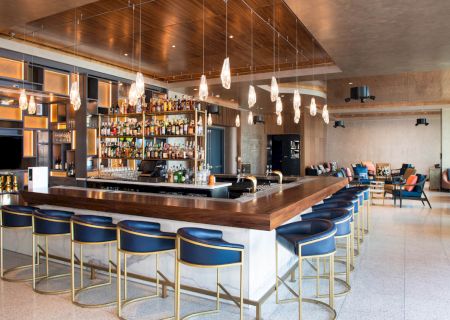 A modern bar with blue chairs, wooden countertops, pendant lights, and a well-stocked back bar in a spacious lounge setting.