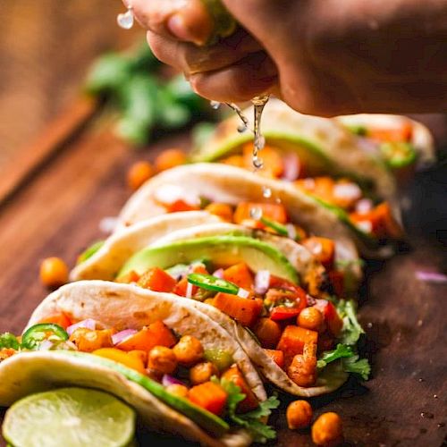 A hand squeezes lime over tacos filled with chickpeas, vegetables, and fresh cilantro, arranged on a wooden board with lime slices.