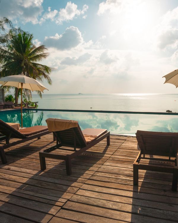 A serene poolside view with lounge chairs, umbrellas, and palm trees, overlooking a calm sea under a partly cloudy sky at sunrise or sunset.
