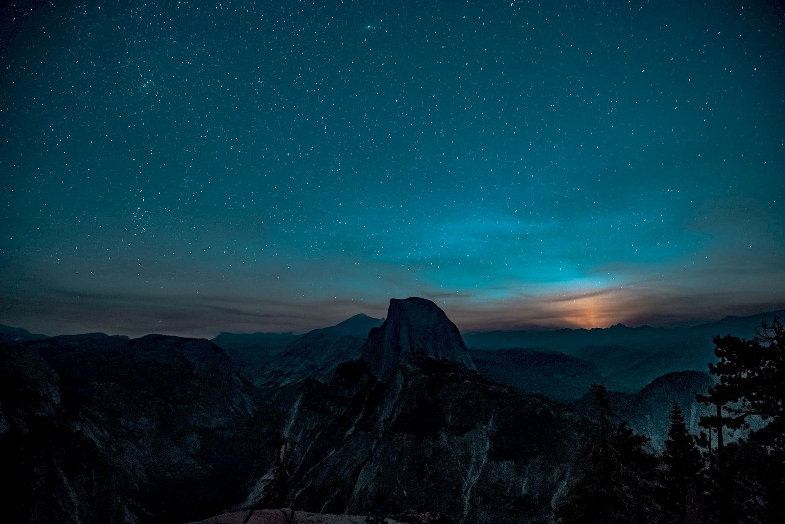 A starry night sky above a mountainous landscape, with a prominent peak silhouetted against a softly glowing horizon.