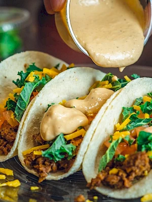 Three tacos filled with ground meat, cheese, lettuce, and topped with sauce are on a plate. Someone is pouring extra sauce on them.
