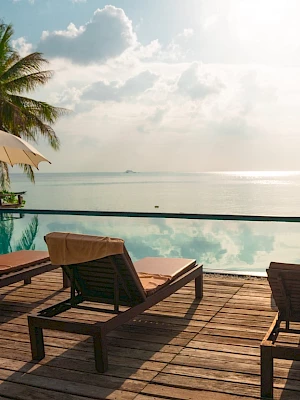 Sunlit beachfront scene with lounge chairs, umbrellas, and palm trees by a pool, overlooking a tranquil sea under a partly cloudy sky.