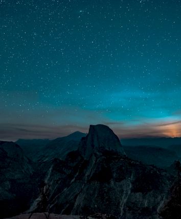 A starry night sky over a mountain silhouette with trees in the foreground, and a glowing light rising behind the peaks.