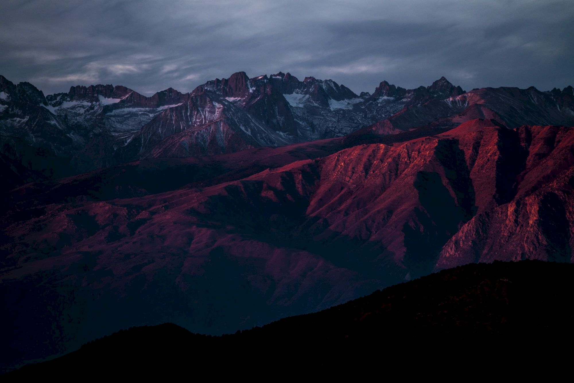 A mountain range is bathed in warm sunset light, with dramatic shadows and a cloudy sky overhead, creating a striking scene.