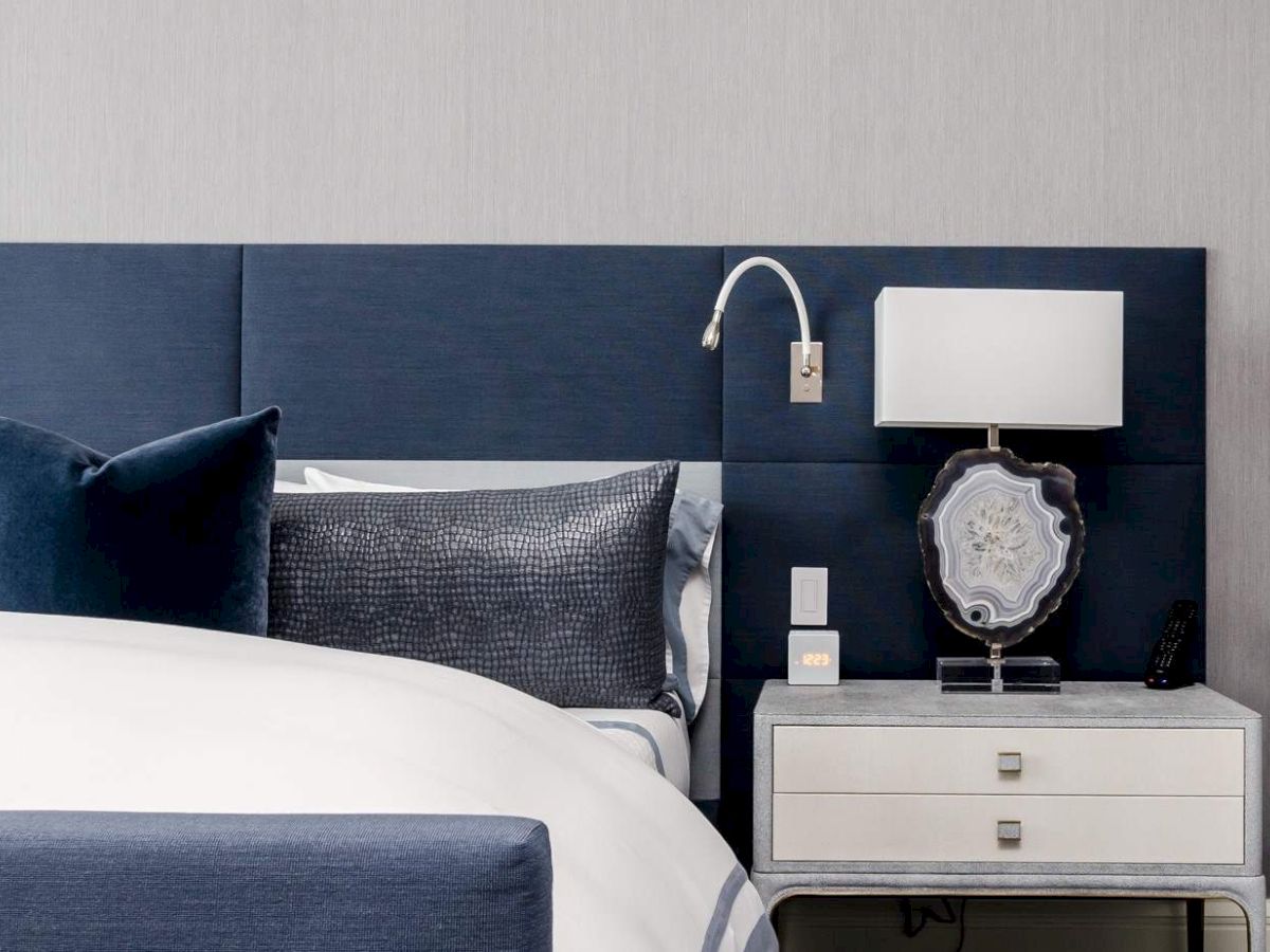 A modern bedroom with a bed and navy headboard, pillows, a nightstand with a lamp, and a reading light attached to the wall.