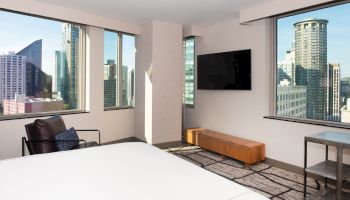 A modern hotel room with a city view features a bed, armchair, wall-mounted TV, and large windows overlooking skyscrapers, adding natural light.