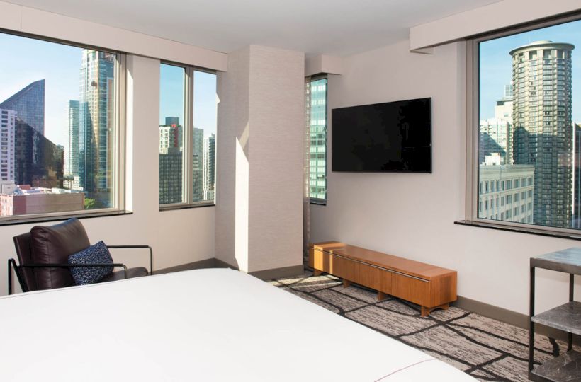 A modern hotel room with a city view features a bed, armchair, wall-mounted TV, and large windows overlooking skyscrapers, adding natural light.