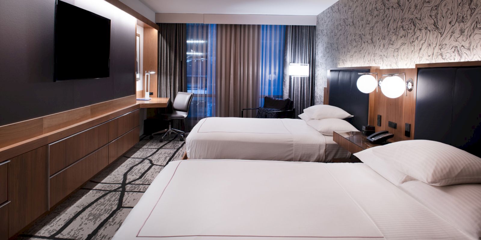The image shows a modern hotel room with two beds, a TV, a work desk, and ambient lighting, featuring a patterned wall.