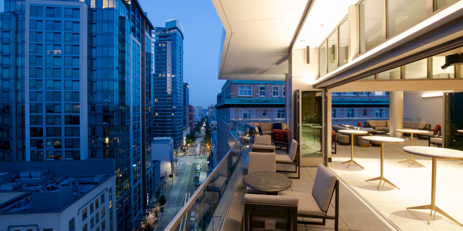 A modern cityscape at dusk featuring tall buildings and a stylish balcony with tables and seating, overlooking a bustling urban street.