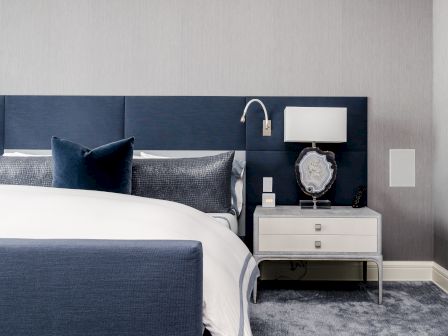 A modern bedroom with a blue upholstered bed, white bedding, navy pillows, and a white nightstand with a lamp and decorative item.