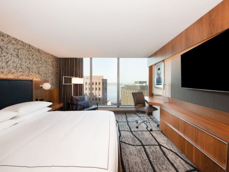 A modern hotel room with a bed, desk, chair, large TV, and window view of buildings, designed with contemporary decor.