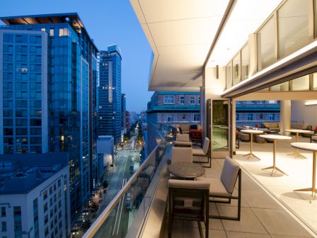 The image shows a modern rooftop balcony at dusk, with city buildings, outdoor seating, and large glass doors opening into an interior space.
