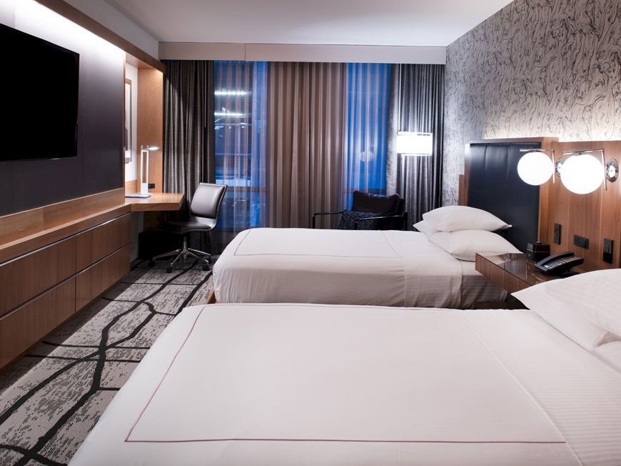 A modern hotel room with two beds, a TV, a desk, and ambient lighting. The decor is sleek and contemporary.