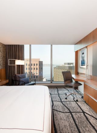 A modern hotel room with a large bed, desk, chair, and wall-mounted TV. The window offers a city view with buildings and water.