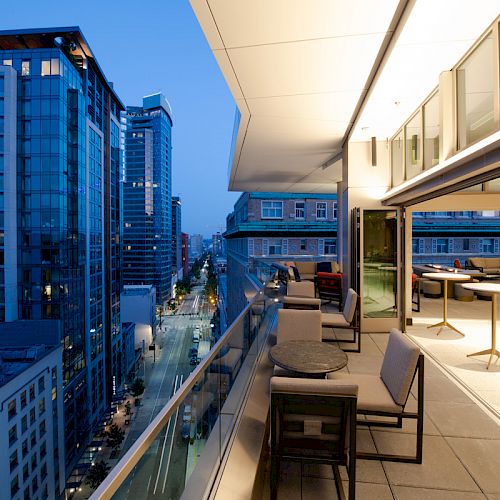A modern balcony at dusk overlooks a city street with tall buildings, featuring seating and outdoor tables near an open indoor area.
