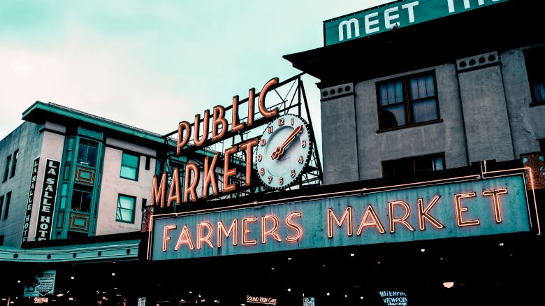 The image shows a famous farmers market entrance with neon signs and a clock, set against a backdrop of buildings and an overcast sky.