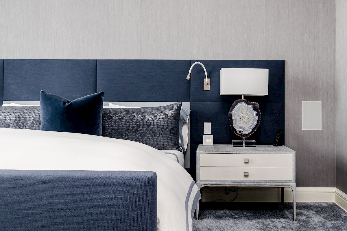 A modern bedroom with a navy upholstered bed, dark pillows, a bedside table with a lamp, and a decorative wall fixture above it.