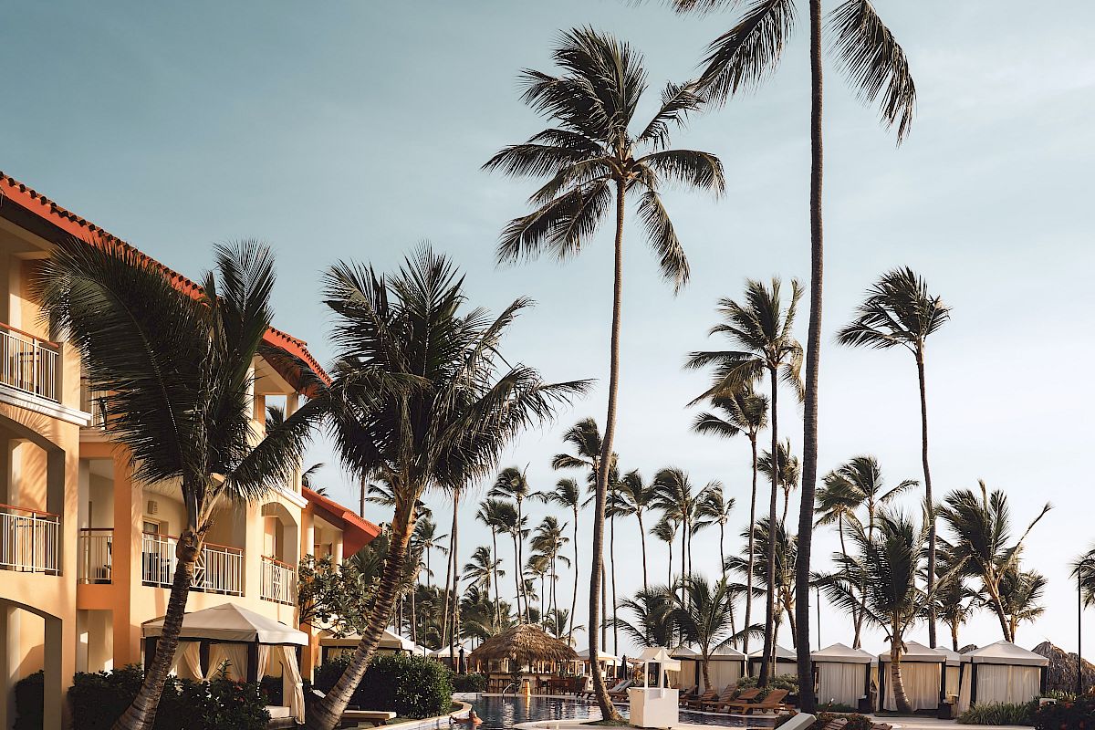 A tropical resort poolside with lounge chairs, palm trees, and a building in the background against a clear sky.