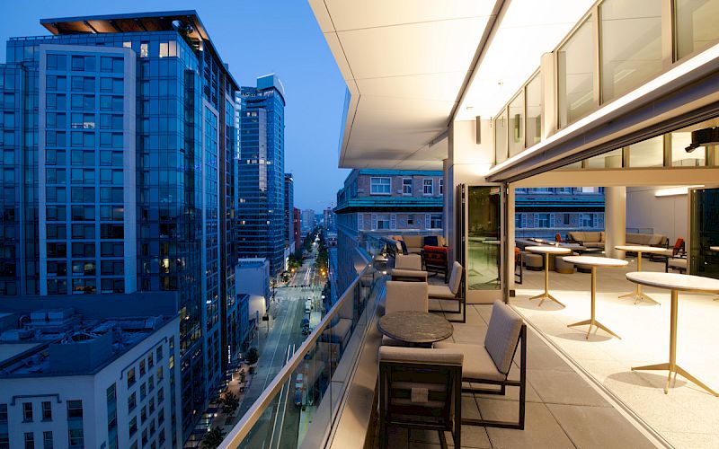 Cityscape at dusk with tall buildings and a glass balcony featuring seating and tables.