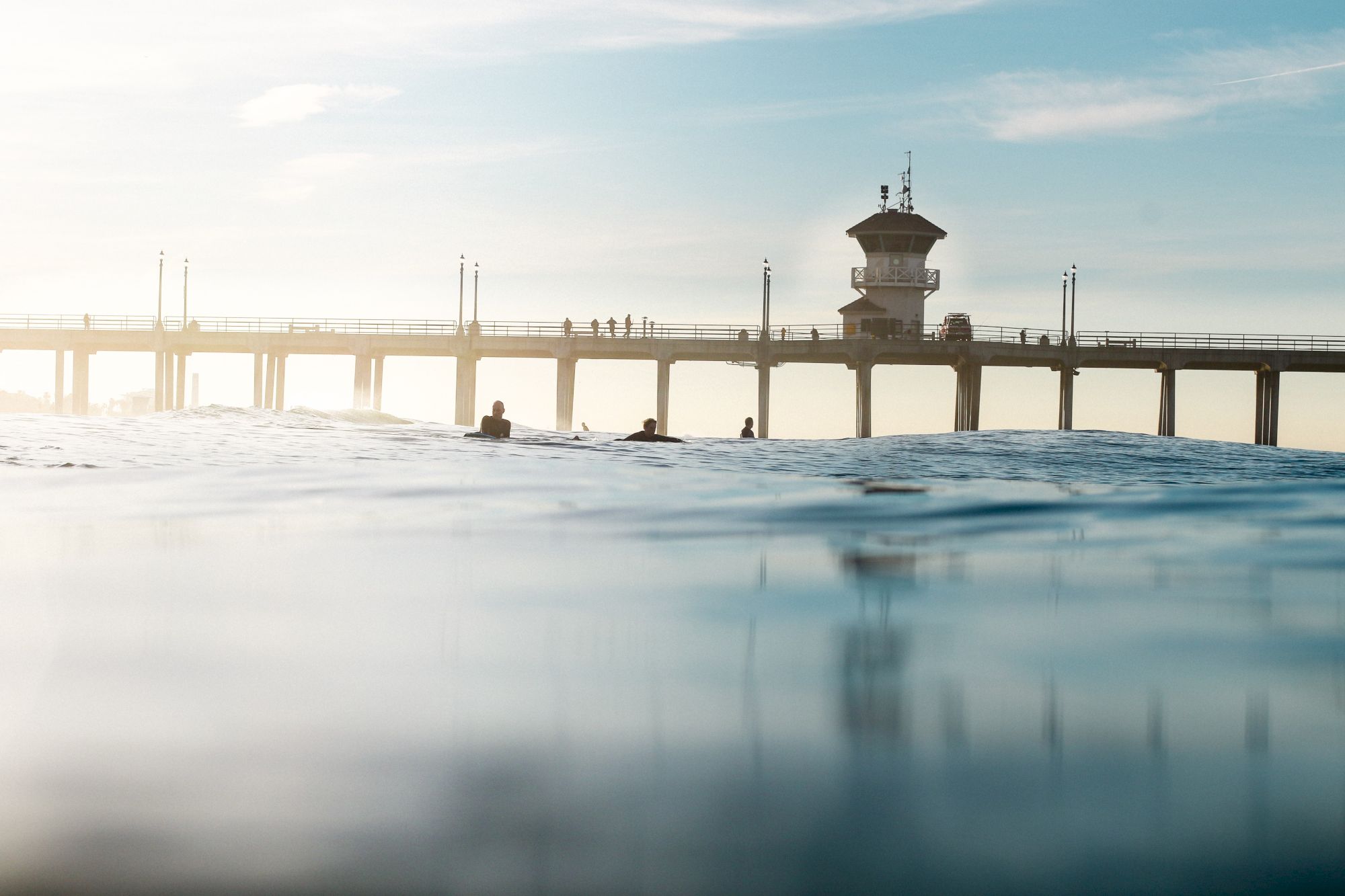 A serene ocean view captures a pier with an observation tower, people strolling, and swimmers in the foreground under clear skies.